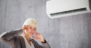 A slightly irritated woman covering her ears with her fingers while standing under a buzzing AC unit.