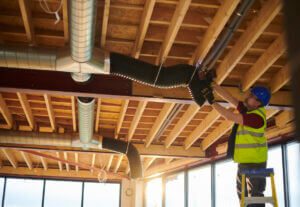 Right-Sized HVAC Ducts: A construction worker in safety gear on a ladder installing HVAC ductwork on an exposed wooden ceiling.